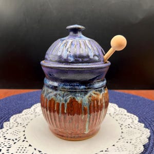 May include: A handmade ceramic jar with a lid and a small wooden spoon. The jar is a mix of colors, including blue, brown, and red. The lid has a ribbed design. The jar is sitting on a white doily.