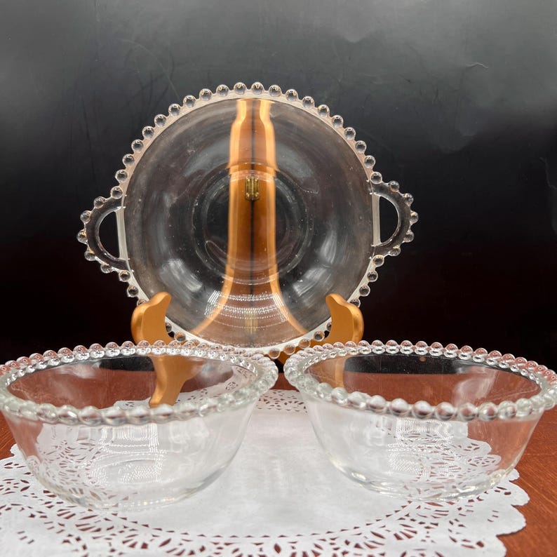 May include: A clear glass serving set featuring a large handled bowl and two smaller bowls. The rims of the bowls are decorated with a beaded design. The set is displayed on a white doily and a wooden stand, suggesting a vintage or antique aesthetic.