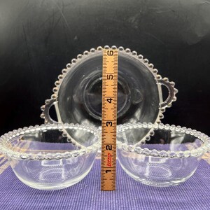 May include: Clear glass serving set featuring a large bowl with handles and two smaller bowls. The rims of the bowls are decorated with a beaded design. The large bowl measures approximately 6 inches in diameter. The small bowls are about 4 inches in diameter.