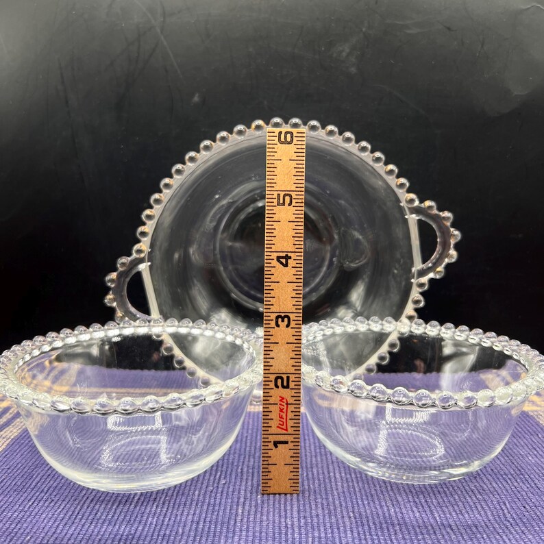 May include: Clear glass serving set featuring a large bowl with handles and two smaller bowls. The rims of the bowls are decorated with a beaded design. The large bowl measures approximately 6 inches in diameter. The small bowls are about 4 inches in diameter.