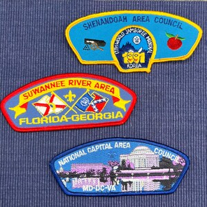 May include: Three embroidered scout patches. The top patch is blue with yellow trim, reading SHENANDOAH AREA COUNCIL and 17TH WORLD JAMBOREE KOREA. The middle patch is blue with red trim, reading SUWANNEE RIVER AREA FLORIDA-GEORGIA. The bottom patch is blue, reading NATIONAL CAPITAL AREA COUNCIL MD-DC-VA with a building illustration.