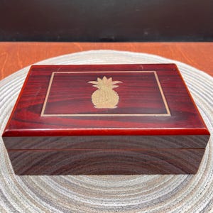 May include: A rectangular dark reddish-brown wooden box with a polished finish. The lid features a centered gold-colored pineapple inlay surrounded by a thin gold-colored rectangular border. The box has a smooth, glossy surface and straight edges.