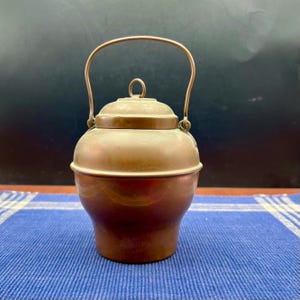 May include: A small, antique copper kettle with a rounded body and a lid. The kettle has a curved handle and a small loop on the lid. The copper has a warm, reddish-brown color. The kettle is sitting on a blue and white striped surface.