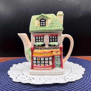 May include: A ceramic teapot shaped like a building with a green shingled roof, a chimney, and a cream-colored facade. The front features the text THE OLD SWEETSHOP, two upper windows with green accents, a storefront window, and a green door with the number 7.