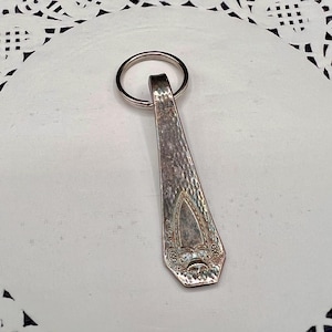 May include: Silver-toned keychain made from a repurposed spoon. The spoon's handle has a hammered texture and decorative floral details. A silver key ring is attached to the top. The keychain is displayed on a white surface.
