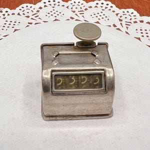 May include: A vintage silver-toned metal tally counter featuring a rectangular display window showing the numbers 5353. The device has a rounded top plunger button and a rectangular base, with a weathered metallic finish and visible signs of age.