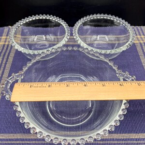 May include: Three clear glass bowls with a beaded rim. The largest bowl has a handle and measures approximately 6 inches in diameter. The smaller bowls are about 4 inches in diameter. The bowls are on a blue and yellow plaid surface.
