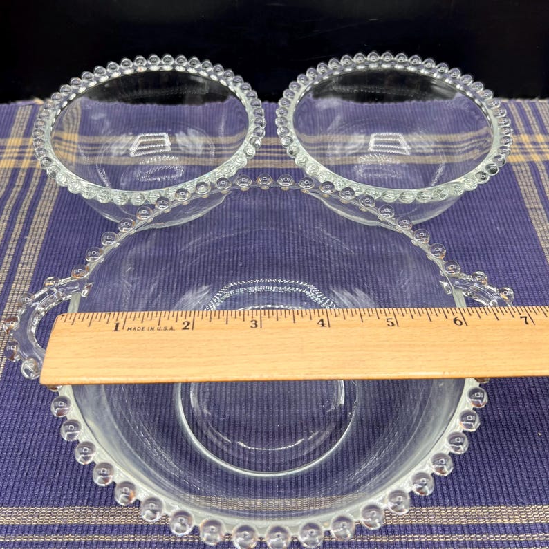 May include: Three clear glass bowls with a beaded rim. The largest bowl has a handle and measures approximately 6 inches in diameter. The smaller bowls are about 4 inches in diameter. The bowls are on a blue and yellow plaid surface.