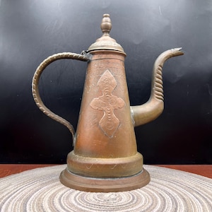 May include: A copper-toned metal pitcher featuring a tapered body, a long curved spout with a textured finish, and a matching handle. The exterior surface is decorated with an embossed cross-shaped motif and a lid topped with a small finial.