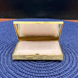May include: Open gold-toned compact with a mirror and a pink powder puff. The compact is rectangular with a hinged lid. The word "Luzier" is printed on the powder puff. The compact is on a blue surface.