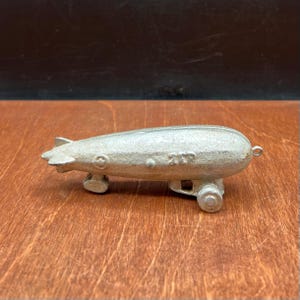 May include: A silver-colored toy zeppelin with the number "212" embossed on the side. The toy has a rounded body, a small tail fin, and two wheels. The zeppelin is made of metal.