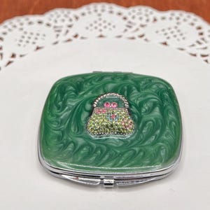 May include: A square, green compact mirror with a swirling pattern. The mirror features a silver frame and a decorative handbag design on the front, embellished with colorful rhinestones. The compact is closed and sits on a white surface.