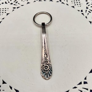 May include: Silverware-themed keychain featuring a spoon design. The spoon has a floral pattern on the handle and a silver key ring. The keychain is made of metal and is a decorative accessory.