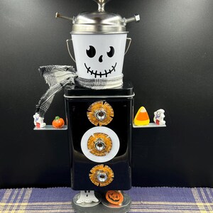 May include: A Halloween-themed metal sculpture resembling a robot. It features a white bucket with a skull face, a black box body with decorative hardware, and small Halloween-themed figurines. A black and white scarf is around the neck.