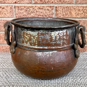 May include: A round copper pot with a wide rim and a weathered, dark brown patina. The pot features two ornate, dark metal side handles with ring attachments. The upper section shows signs of worn silver-toned plating over the copper surface.