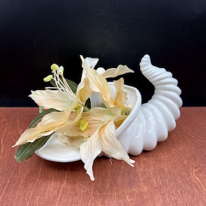 May include: A white ceramic cornucopia vase filled with peach-colored flowers and green leaves. The vase has a curved, horn-like shape with a textured, ribbed exterior. The flowers have yellow centers and are arranged to spill out of the vase.