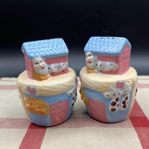 May include: A pair of ceramic salt and pepper shakers shaped like a house with animals peeking out. The shakers are painted in shades of pink, blue, and yellow with brown accents.