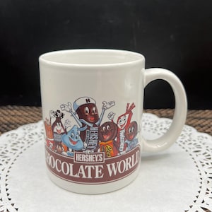 May include: White ceramic mug featuring Hershey's Chocolate World characters. The mug has a brown band with the words "Hershey's Chocolate World" printed on it. The mug has a white handle and is on a white doily.