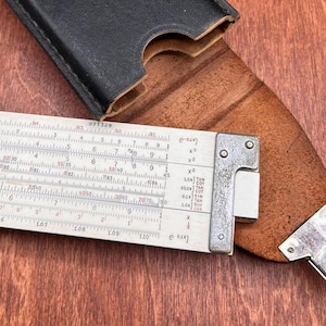 May include: A vintage slide rule with a metal and white scale, partially visible, with numbers and markings. The slide rule is partially inside a black leather case, with a brown leather backing.