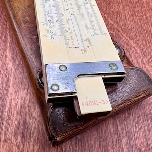 May include: A vintage slide rule with a cream-colored scale and metal frame, resting on a brown leather case. The slide rule features various numerical markings and a small rectangular piece with the text "<4091-3>".