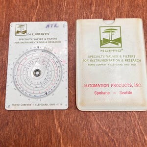 May include: A white Nupro circular slide rule calculator with a clear rotating cursor, featuring printed scales and the text Nupro Specialty Valves & Filters for Instrumentation & Research, alongside a matching white protective sleeve with red text.