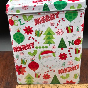 May include: A rectangular Christmas tin with a white background, adorned with festive red, green, and white holiday graphics. The design includes Christmas trees, ornaments, holly, candy canes, gift boxes, snowflakes, and the words "MERRY" and "NOEL". A Santa Claus graphic is also featured.