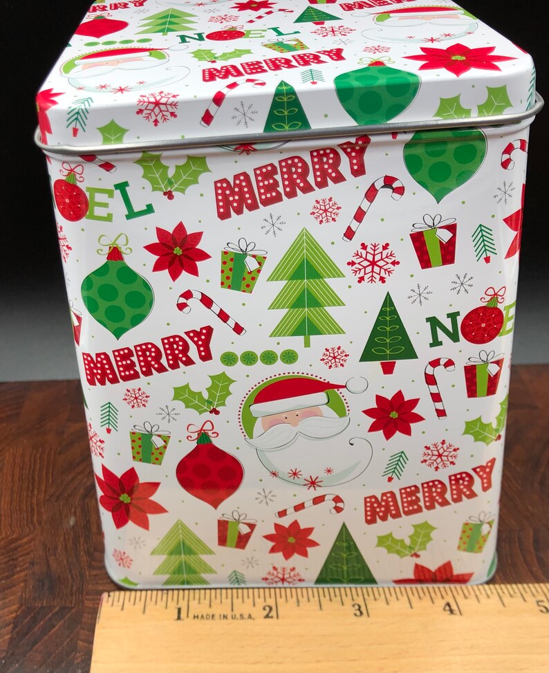 May include: A rectangular Christmas tin with a white background, adorned with festive red, green, and white holiday graphics. The design includes Christmas trees, ornaments, holly, candy canes, gift boxes, snowflakes, and the words "MERRY" and "NOEL". A Santa Claus graphic is also featured.