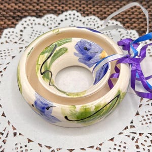 May include: A round, cream-colored ceramic ornament with a central hole, hand-painted with blue floral motifs and green leaves, featuring black line accents and a decorative purple and blue ribbon tied to the side for hanging purposes.
