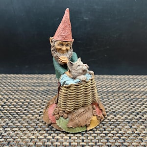 May include: A whimsical figurine depicting a gnome and a cat. The gnome, wearing a red pointed hat and green jacket, is smiling at the cat in a woven basket. The base features the text "CATFISHING".
