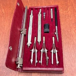 May include: A vintage drafting instrument set housed in a red velvet-lined case. The collection includes several silver-toned metal compasses, a long beam compass bar, a small black ink container, and a black-handled technical drawing pen.