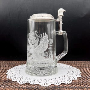 May include: A clear glass beer stein featuring an etched design of an eagle in flight with mountains and clouds. The stein has a metal lid with a sculpted eagle head thumb lift and a sturdy glass handle, providing a classic look for beverage service.