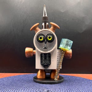 May include: A robot-shaped sculpture constructed from mixed objects, featuring a Polaroid flash unit face with yellow eyes, a metal bottle stopper head, copper-toned limbs, and a blue cube accessory held by a spring-loaded arm.