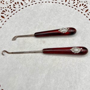 May include: Two vintage crochet hooks. Each hook has a dark red handle with a silver-colored decorative accent. One hook has a curved end, and the other has a flat, angled end. The hooks are displayed on a white surface.