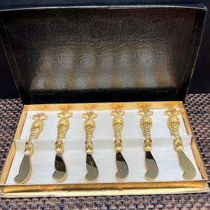 May include: A set of six gold-colored butter knives in a black and gold box. Each knife features a pineapple design on the handle and a curved blade. The knives are arranged neatly in a white insert within the box.