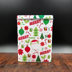 May include: Rectangular white Christmas tin with a festive pattern. The design includes red and green ornaments, Christmas trees, candy canes, and the word "MERRY" in red. A Santa Claus graphic is also featured.