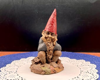 Könnte beinhalten: Eine kleine Gartenzwerg-Figur mit einer spitzen roten Mütze, einem langen, strukturierten Bart und einer blauen Jacke. Der Zwerg sitzt in einer ruhenden Haltung mit den Händen am Gesicht auf einem Sockel aus modellierten Herbstblättern.