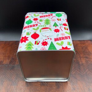 May include: A rectangular metal Christmas tin with a festive design. The white lid features red, green, and white holiday graphics, including Santa Claus, ornaments, Christmas trees, and the word "MERRY". The sides are a metallic brown color.