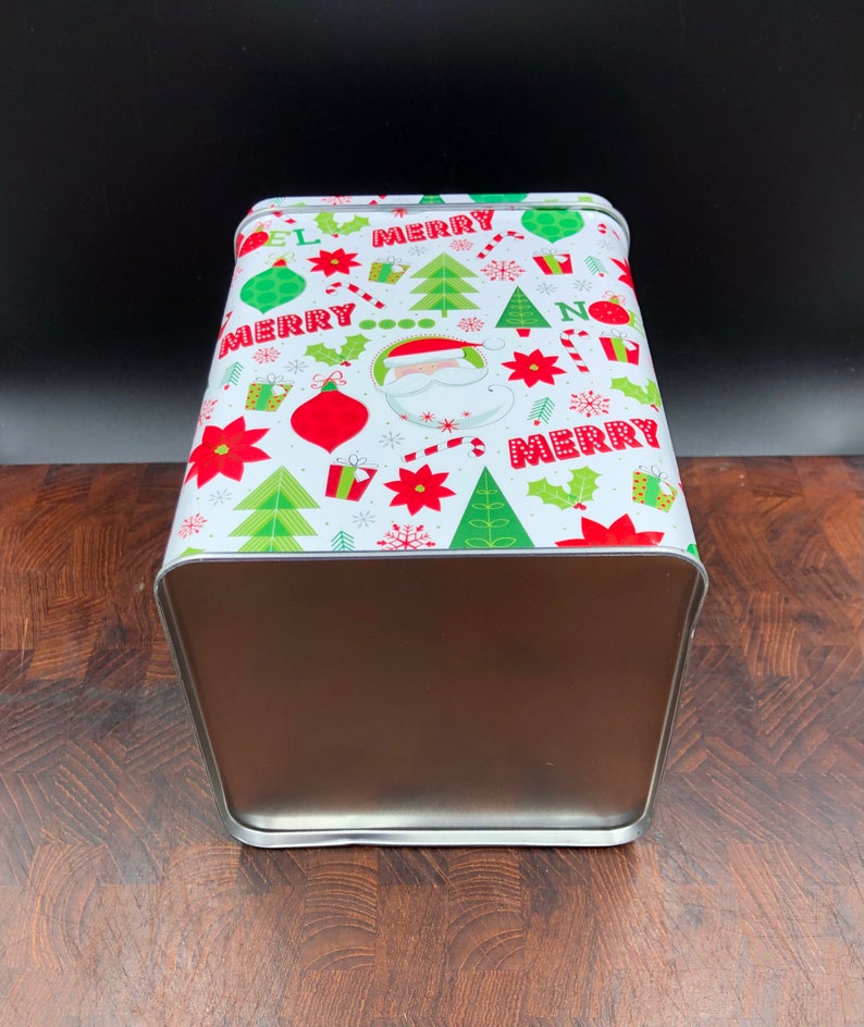 May include: A rectangular metal Christmas tin with a festive design. The white lid features red, green, and white holiday graphics, including Santa Claus, ornaments, Christmas trees, and the word "MERRY". The sides are a metallic brown color.