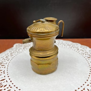 May include: A vintage brass oil lamp with a handle and a small spout. The lamp has a round base and a domed top with etched details. It is sitting on a white doily.
