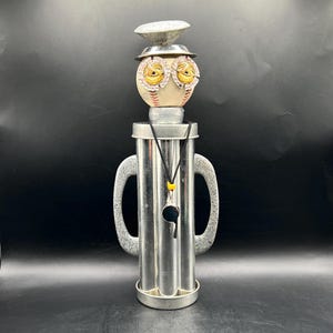 May include: A decorative figurine constructed from a metallic cylinder with two speckled gray handles, topped with a baseball head featuring bottle cap eyes and a metal hat. A black cord necklace with a small black whistle hangs around the neck.