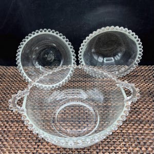 May include: Three clear glass bowls with beaded rims. Two bowls are round, and one is oval with handles. The bowls are made of clear glass and have a delicate, vintage appearance. The bowls are arranged on a brown surface.