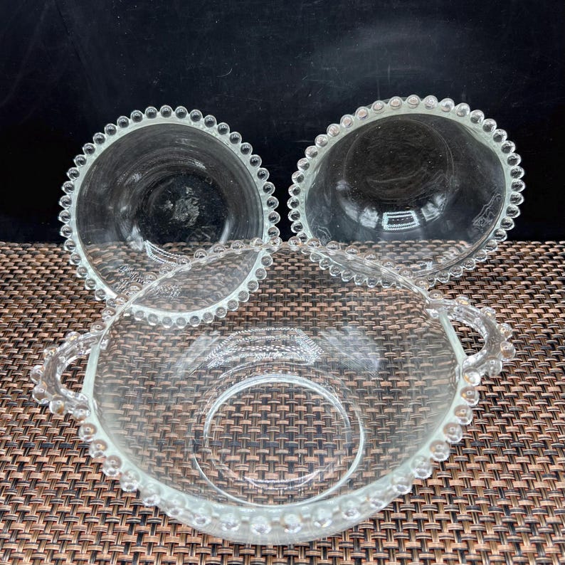 May include: Three clear glass bowls with beaded rims. Two bowls are round, and one is oval with handles. The bowls are made of clear glass and have a delicate, vintage appearance. The bowls are arranged on a brown surface.