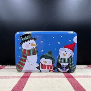 May include: A rectangular blue tin featuring a snowman and two penguins, all wearing winter accessories. The snowman has a black top hat and a striped scarf. The penguins wear hats and scarves, one holding a gift. White snowflakes are scattered across the blue background.