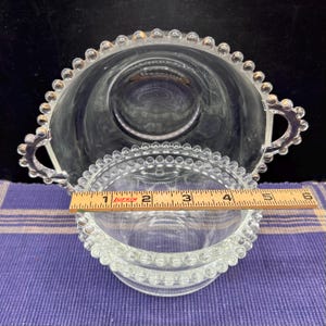 May include: Clear glass serving bowl and small dish with a beaded rim. The bowl has two handles and is approximately 5 inches in diameter. The small dish is about 4 inches in diameter. Both are on a purple and white checkered surface.