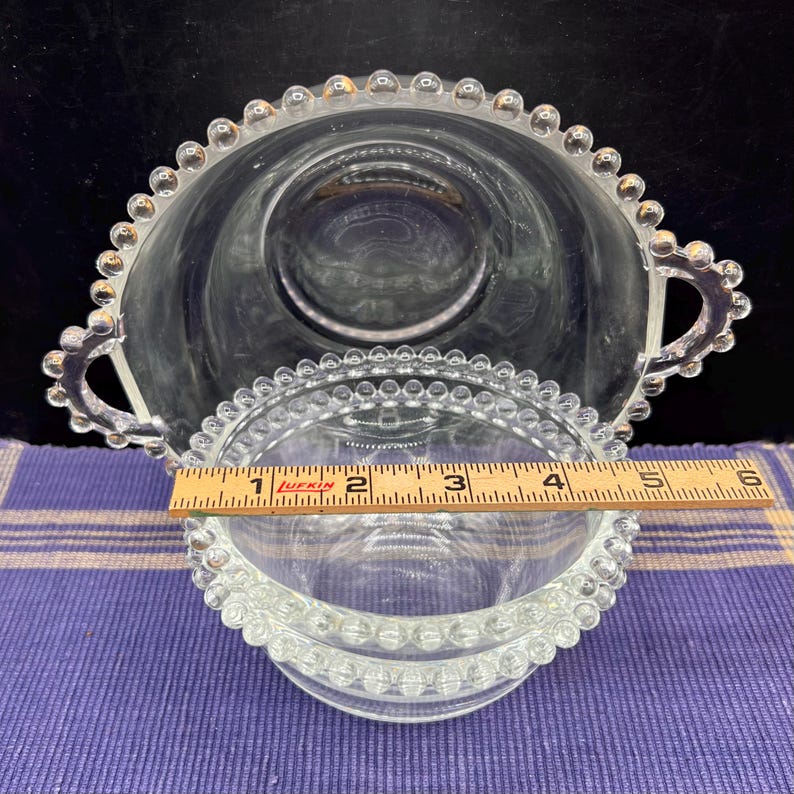 May include: Clear glass serving bowl and small dish with a beaded rim. The bowl has two handles and is approximately 5 inches in diameter. The small dish is about 4 inches in diameter. Both are on a purple and white checkered surface.