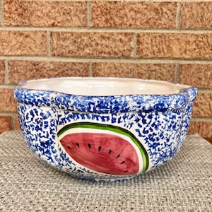 May include: A ceramic bowl featuring a blue and white sponge-painted exterior pattern. The front of the bowl displays a hand-painted watermelon slice graphic with a pink interior, black seeds, and a green rind against the speckled blue background.