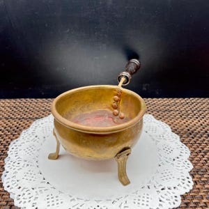 May include: A small, antique brass pot with three decorative legs. The pot has a wooden handle and a series of small, round beads attached to the inside. The pot sits on a white, lacy doily.