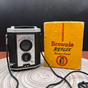 May include: A black and silver Eastman Kodak Brownie Reflex Synchro Model camera with a black neck strap, positioned next to its original yellow cardboard box featuring the text Brownie REFLEX Synchro Model and a graphic illustration of the camera.