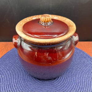 May include: A ceramic casserole dish with a lid. The dish is a deep burgundy color with a lighter brown rim and lid. It has two side handles and a knob on the lid. The dish sits on a blue placemat.