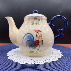 May include: Cream-colored ceramic teapot with a blue handle and lid. The teapot features a rooster illustration with blue, red, and yellow accents. The teapot is sitting on a white lace doily.